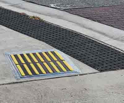 Image of a intersection corner.  Close up of Tactile Direction Indicator (2’x2’ square with raised yellow and black stripes) placed parallel to traffic to indicate the correct alignment.  The TDI is placed on the sidewalk in front of a blended, rounded corner with black truncated domes.